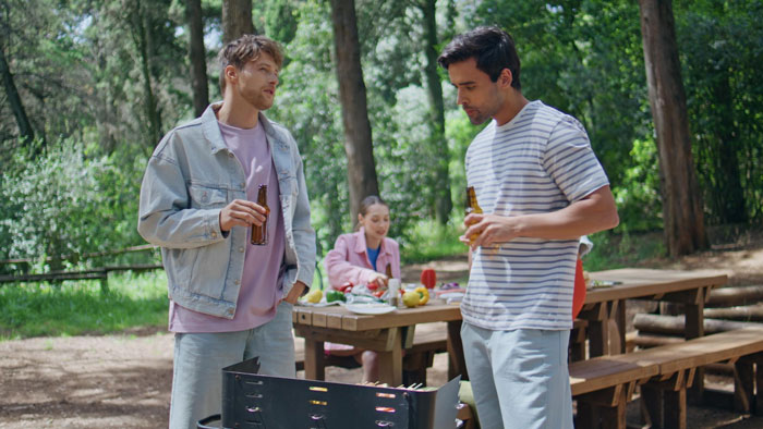 Two men holding beer bottles near a grill in a park, while a woman sits at a picnic table in the background. - 10