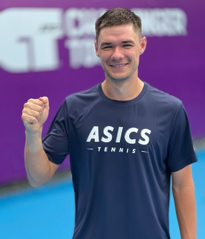 Kamil Majchrzak smiling and raising fist wearing ASICS tennis shirt, related to viral US Open hat snatching incident.