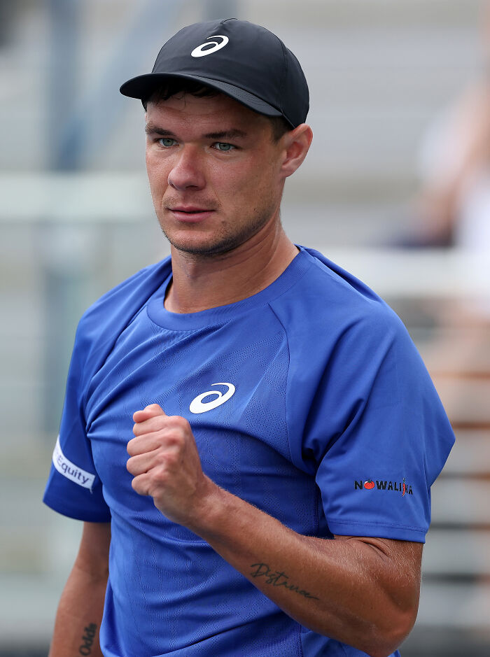 Kamil Majchrzak wearing a blue shirt and black cap, clenching fist during a tennis match at the US Open event.