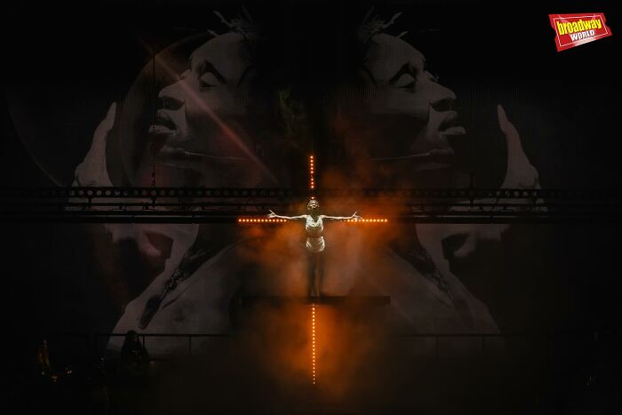 Cynthia Erivo with long nails portrayed as Jesus in a dramatic stage scene with fiery cross and dark background. Cynthia Erivo with long nails portrayed as Jesus in a dramatic stage scene with fiery cross and dark background.