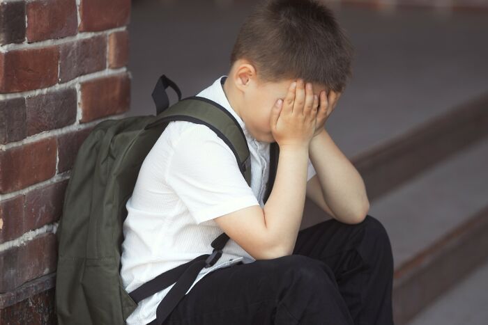 Young boy with backpack sitting on steps covering his face, portraying feelings of rejection and emotional distress.