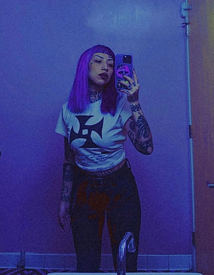 Young woman with purple hair and tattoos taking a mirror selfie in a dimly lit room, symbolizing school shooting planning concerns - 2
