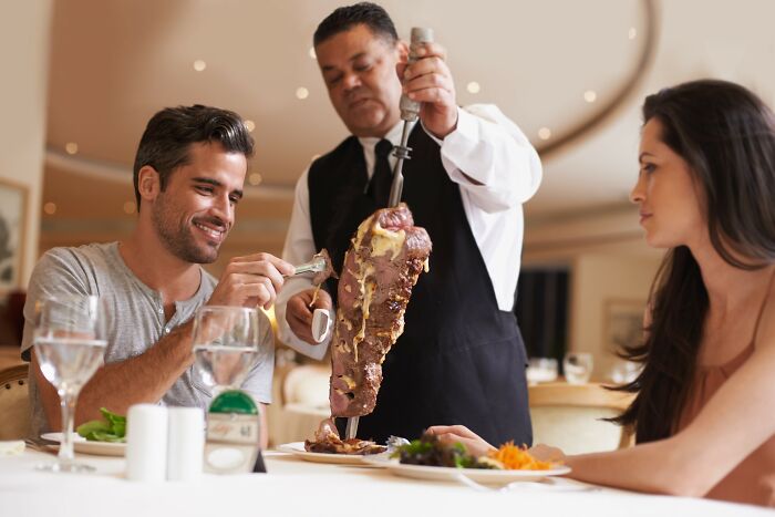 Waiter carving meat tableside for a couple dining in a restaurant, showing a moment restaurant workers deserved a bonus.