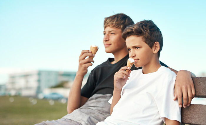 Two boys sitting on a bench eating ice cream, enjoying a moment that evokes childhood memories sweet but unsettling in retrospect.