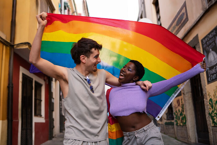 Two friends joyfully holding a rainbow flag together on a street, evoking nostalgic childhood memories that feel unsettling now.