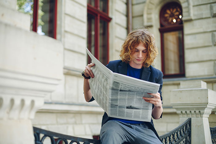 Young man with curly hair reading a newspaper outdoors, reflecting on hot take opinions related to boomers.
