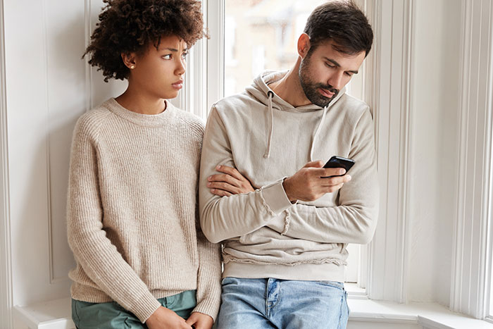 Young man and woman standing by window, discussing a hot take while using a smartphone, reflecting generational perspectives.