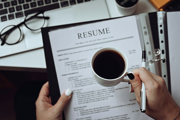 Person holding a resume and coffee cup, preparing to write with a pen near a laptop and glasses on a desk.