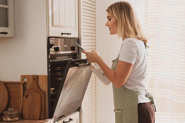 Woman in apron checking oven, using smartphone, illustrating a hot take related to boomers and cooking habits.