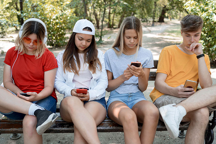 Four teenagers sitting on a bench outdoors, each focused on their smartphone, reflecting hot take discussions with boomers.