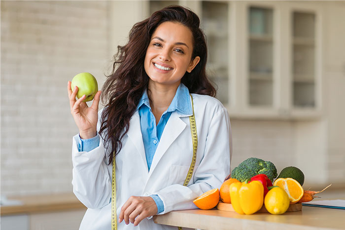 Woman nutritionist holding an apple and smiling with fresh fruits and vegetables, related to pseudoscience and scams.