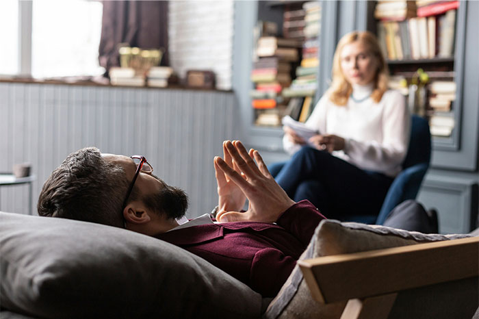 Man lying on a couch talking with a female therapist in a book-filled office discussing pseudoscience and scams.