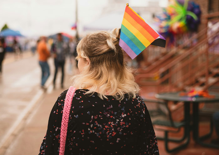 Person with shoulder-length hair holding a rainbow pride flag walking outdoors among a casual crowd, main SEO keyword marriage.