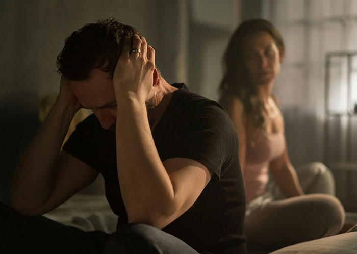 Man holding his head in distress while a woman sits behind him, highlighting marriage challenges and unexpected issues.