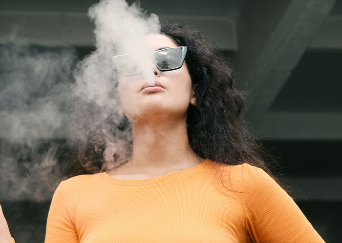 Woman wearing sunglasses and an orange top exhaling smoke, representing things not mentioned until after marriage.