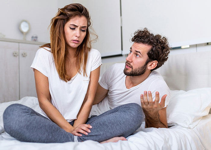 Couple sitting on bed having a serious conversation about things not mentioned until after marriage, showing concern and confusion.