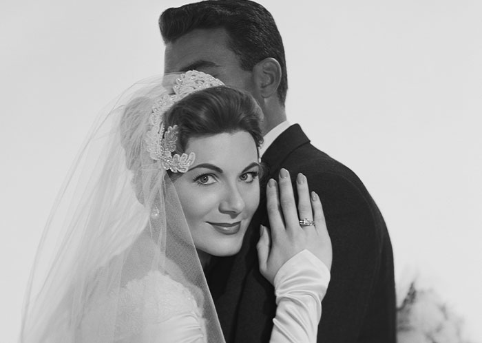 Bride wearing a veil and wedding dress showing her ring while embracing groom, illustrating things not mentioned until after marriage.