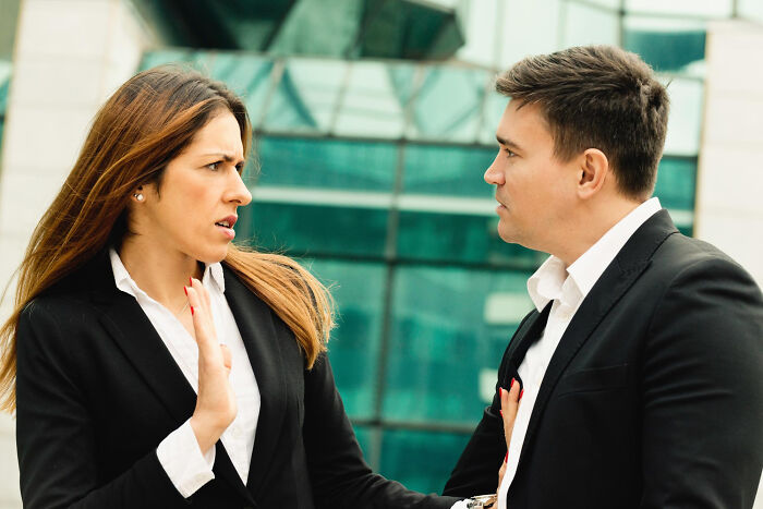 Woman stopping man with hand raised, both in business attire, outside modern glass building, ticking big lifetime event concept.