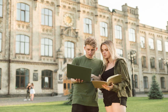 Two students discussing notes on a laptop and book outside a historic university building ticking big lifetime event.