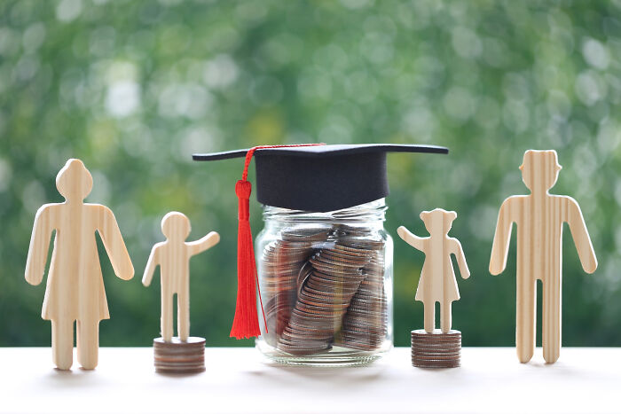 Wooden family figures stand beside a jar of coins with a graduation cap, symbolizing ticking big lifetime event savings.