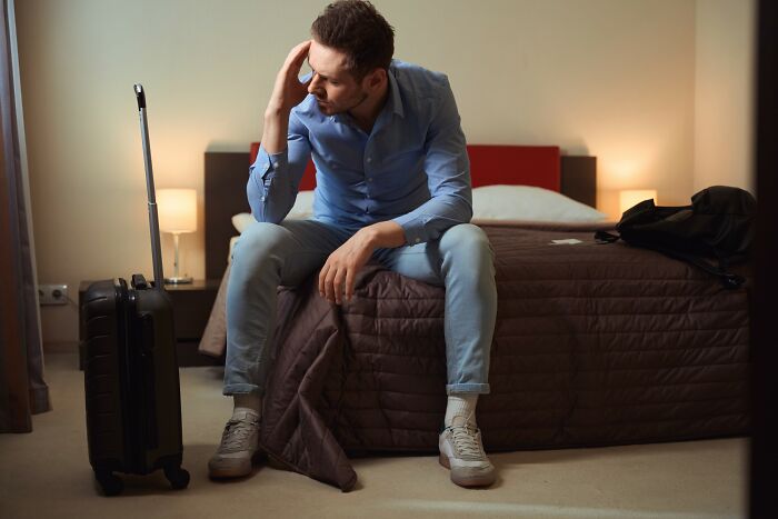 Man sitting on bed with luggage nearby, looking upset after a brutal rejection in a personal relationship story.