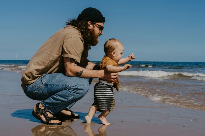 Man with tattoos and hat helping baby walk on beach illustrating gender-based double standards concept.