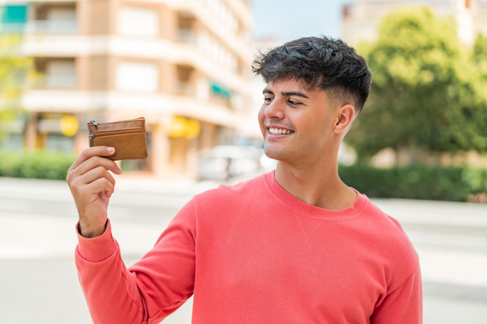 Young man smiling and holding a wallet outdoors, illustrating common tourist traps related to money and spending.