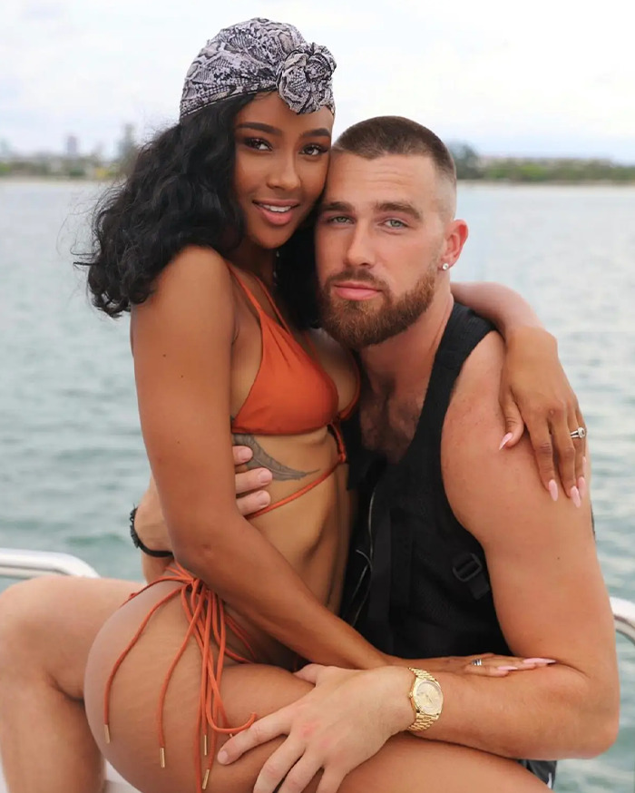 Travis Kelce's ex wearing a headscarf and bikini, embracing a man with a beard while sitting by the water.
