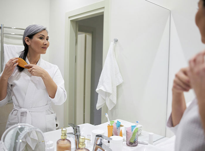Middle-aged woman brushing hair in bathroom mirror, reflecting spooky encounters that still haunt people today.