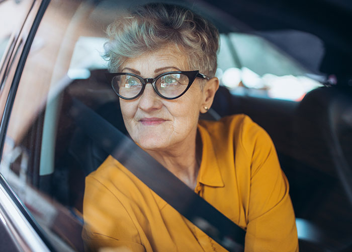Older woman with gray hair and glasses wearing a mustard shirt sitting in a car, representing unhinged MILs sayings.