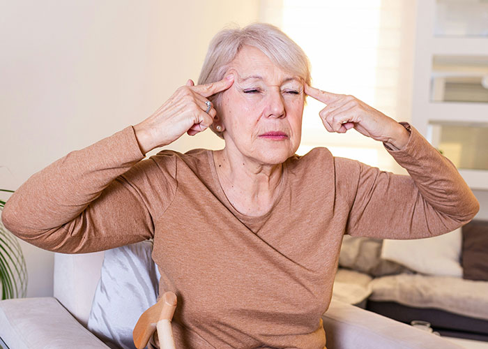 Elderly woman frustrated, sitting indoors, expressing stress with fingers on temples, illustrating unhinged MILs comments.