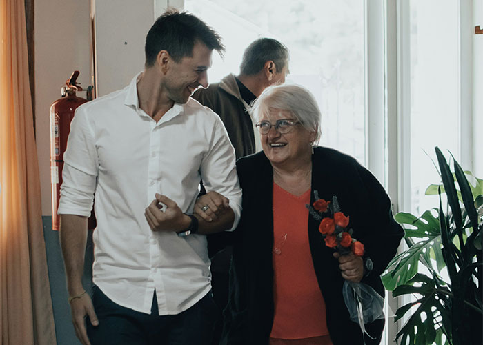Young man smiling with elderly woman holding flowers, illustrating unhinged MILs saying inappropriate things out loud.