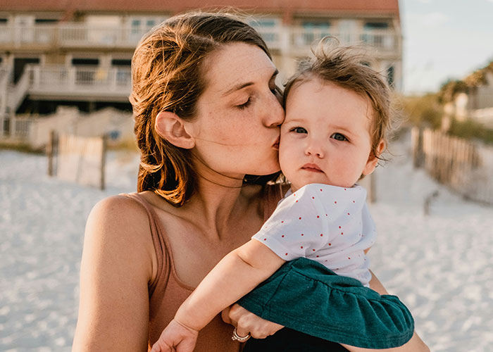 Mother kissing her baby at the beach, capturing tender moments amidst unhinged things MILs said out loud.