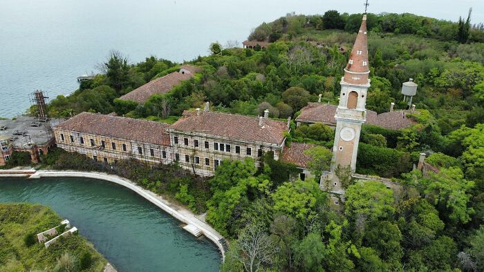 Aerial view of an abandoned building and a tall clock tower surrounded by dense greenery near water.