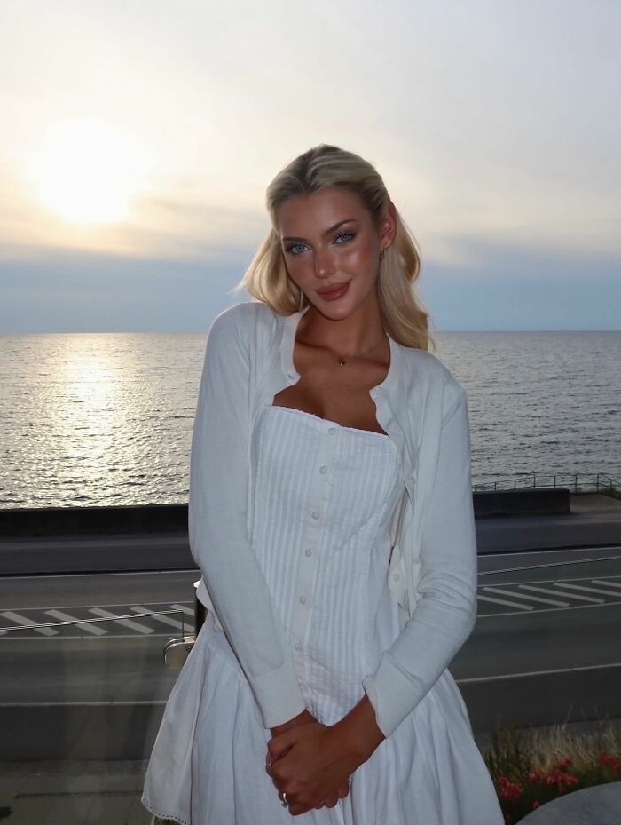 Blonde woman posing by the ocean at sunset, wearing a white dress and cardigan, beauty captured in Miss Universe winners photos