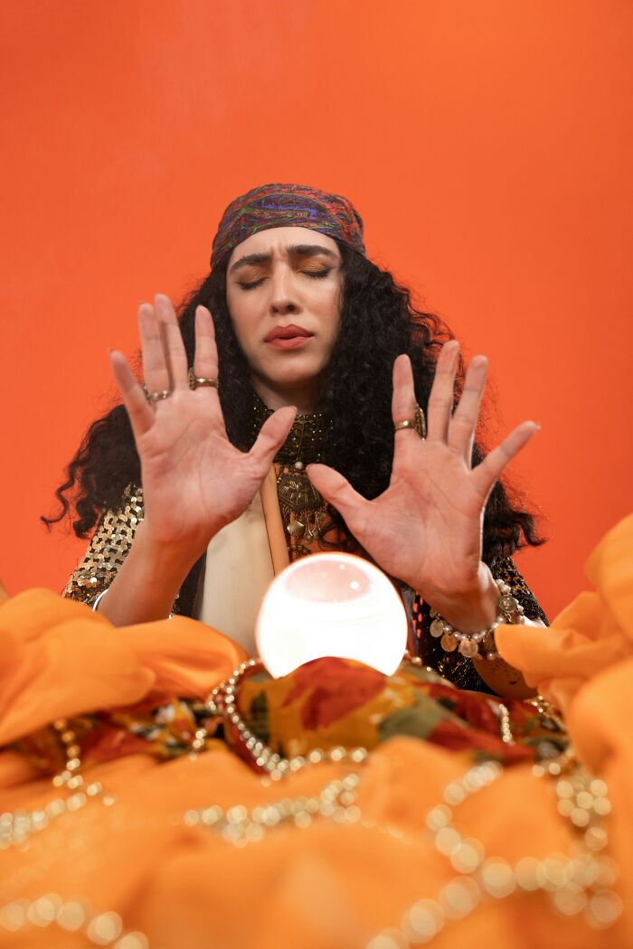 A psychic woman with eyes closed and hands raised over a glowing crystal ball, surrounded by orange fabric.