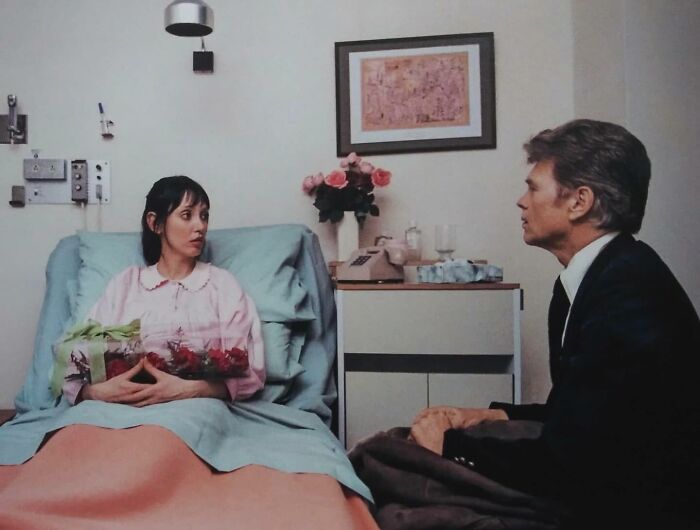 Woman in vintage hospital gown holding flowers, talking with a man in a suit in a nostalgic June Cleavers vintage closet scene