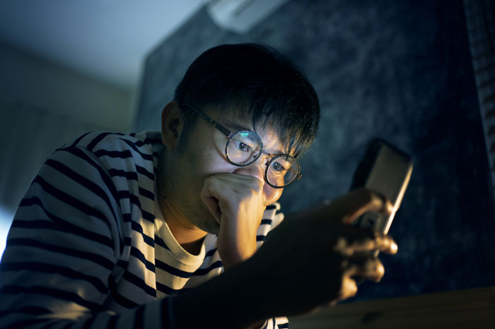 Young man wearing glasses and striped shirt, deeply focused on his phone in a dark room, reflecting water sign emotions.