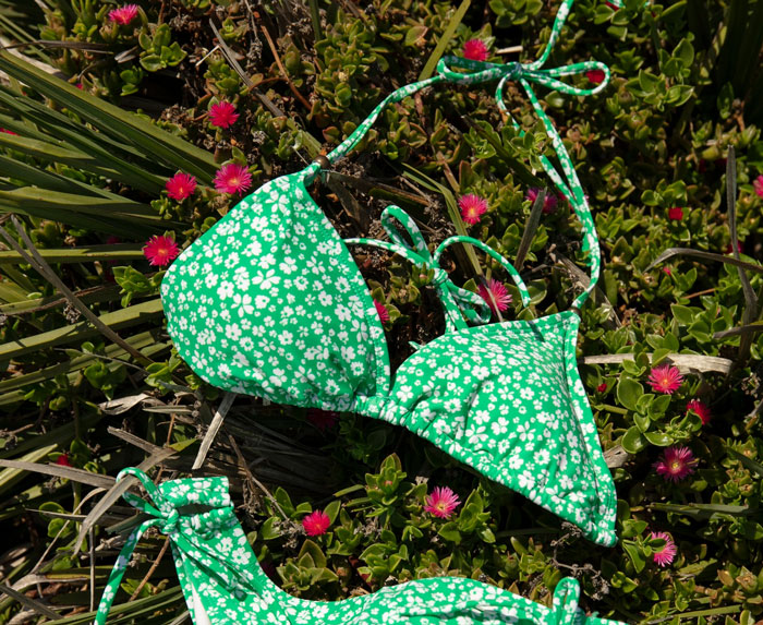 Green floral bikini set laid on vibrant plants with small pink flowers in a sunny own yard setting.