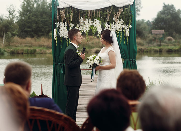 Bride and groom standing under floral arch by the lake during outdoor wedding to avoid nightmare ex at family event - 1