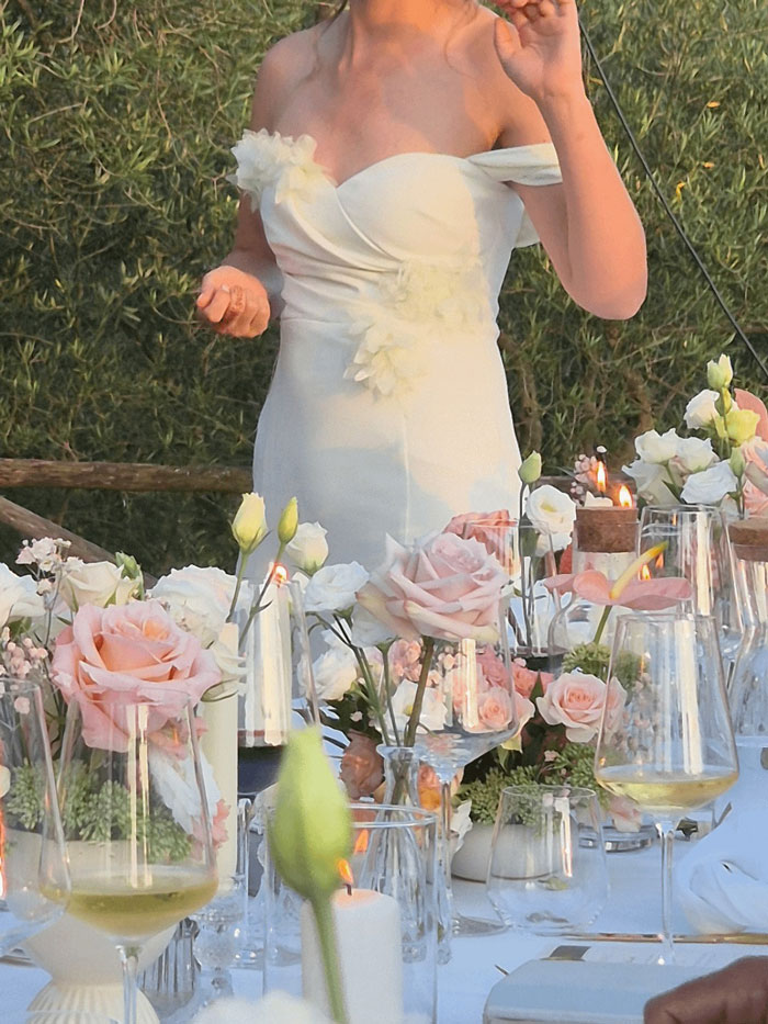 Woman wearing white wedding dress at outdoor wedding table decorated with pink roses and lit candles.