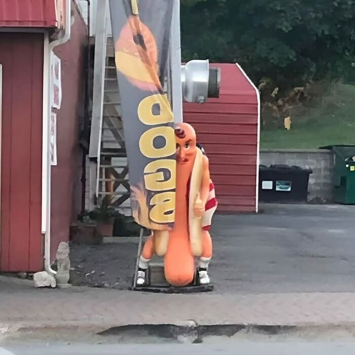 Cursed image of a person in a hot dog costume hiding behind a food banner outside a building.