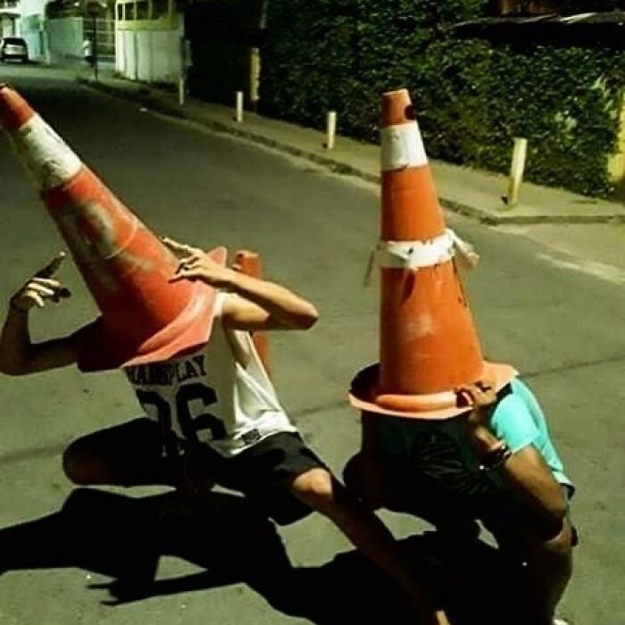 Two people crouching on a street with large traffic cones covering their heads in a cursed image style.