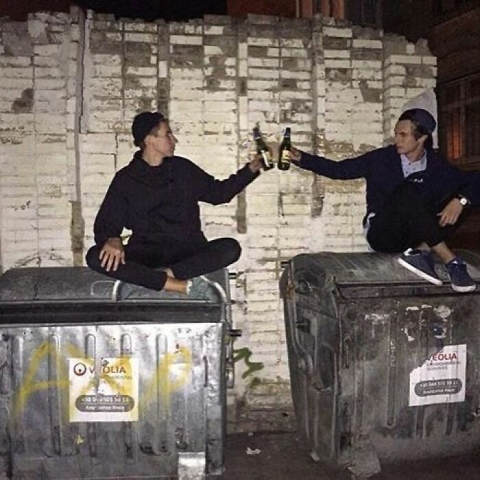 Two young men sitting on dumpsters at night, clinking beer bottles in a scene of daily cursed images on Instagram.