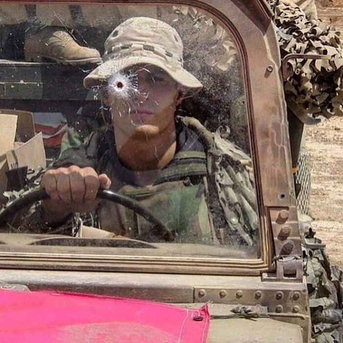 Soldier driving a military vehicle with a bullet hole on the windshield captured in a cursed image.