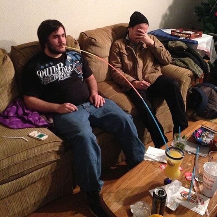 Two men on a couch with a long straw reaching from one man’s mouth to a drink on the table, a cursed image from IG page.