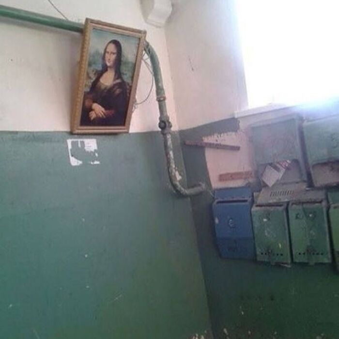 Framed Mona Lisa portrait oddly hanging on a wall with exposed pipes in a worn-down room, cursed images from IG page.