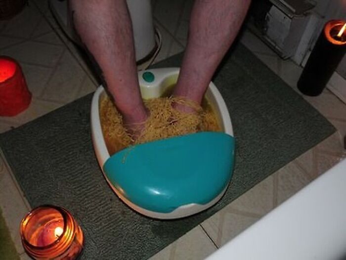 Feet soaking in a foot spa filled with cooked spaghetti noodles, surrounded by lit candles in a bathroom setting.