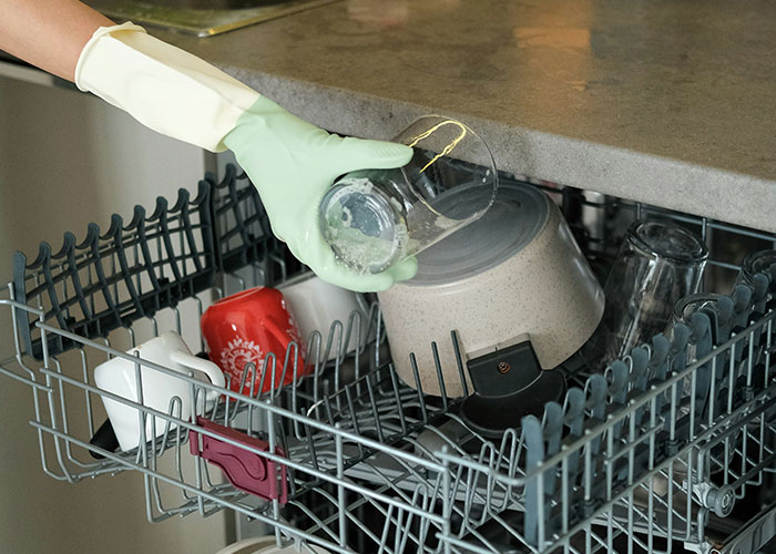 Person wearing green gloves loading a dishwasher, cleaning glasses and kitchen items as part of family routines assumed by many.