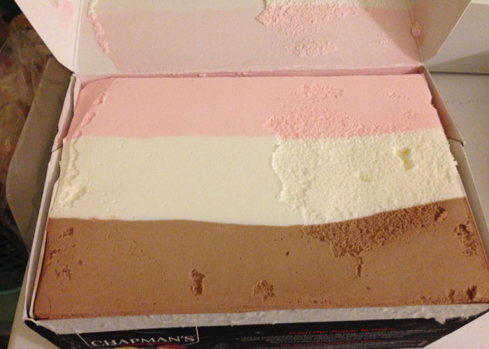 Partially eaten Neapolitan ice cream in a box, showing layers of chocolate, vanilla, and strawberry flavors in a family setting.